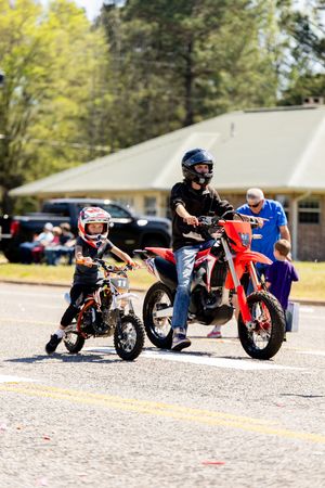 Adult and child riding red dirt bikes in helmets during an outdoor parade on a sunny day.