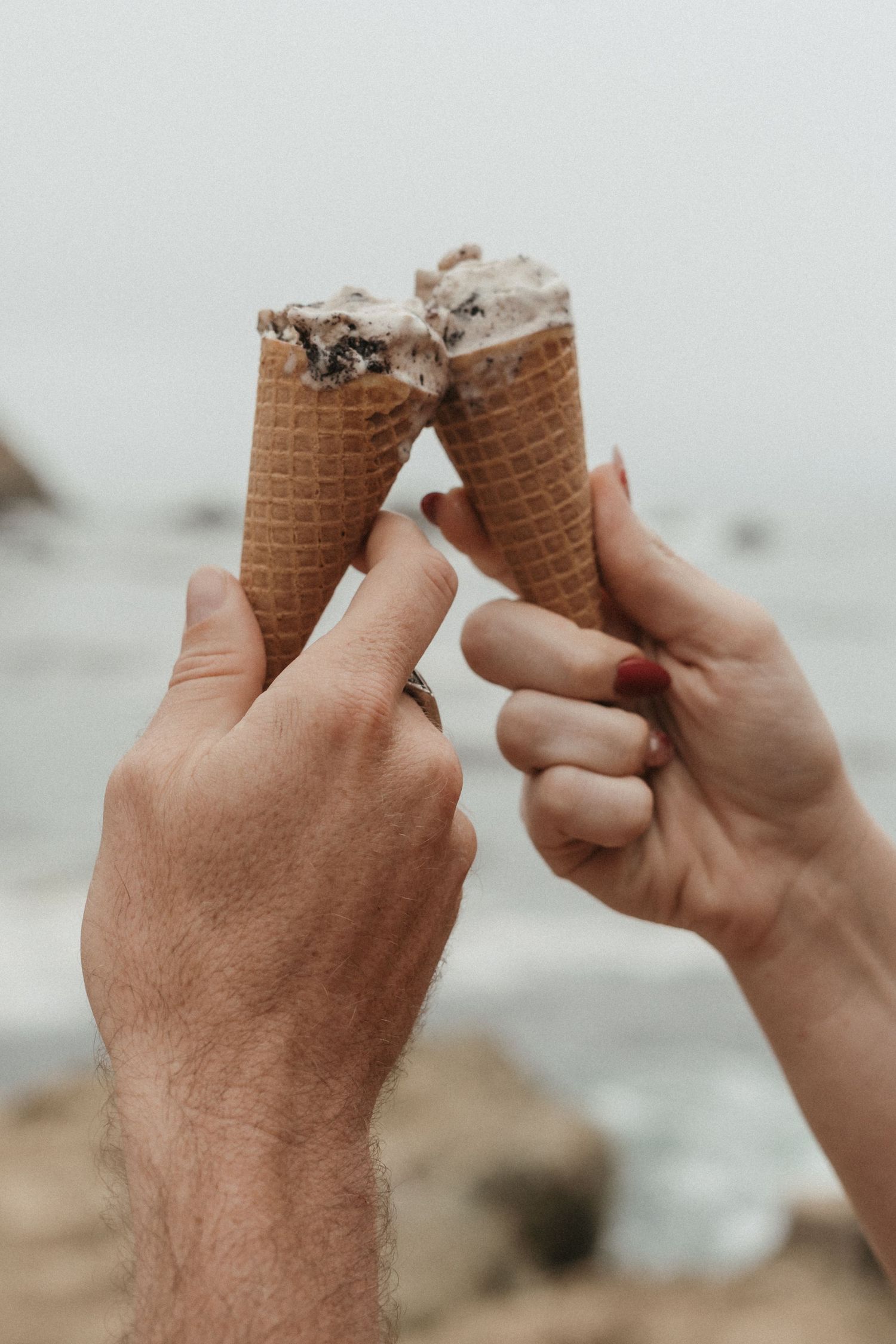 Two hands clinking waffle ice cream cones together at the beach on a cloudy day.