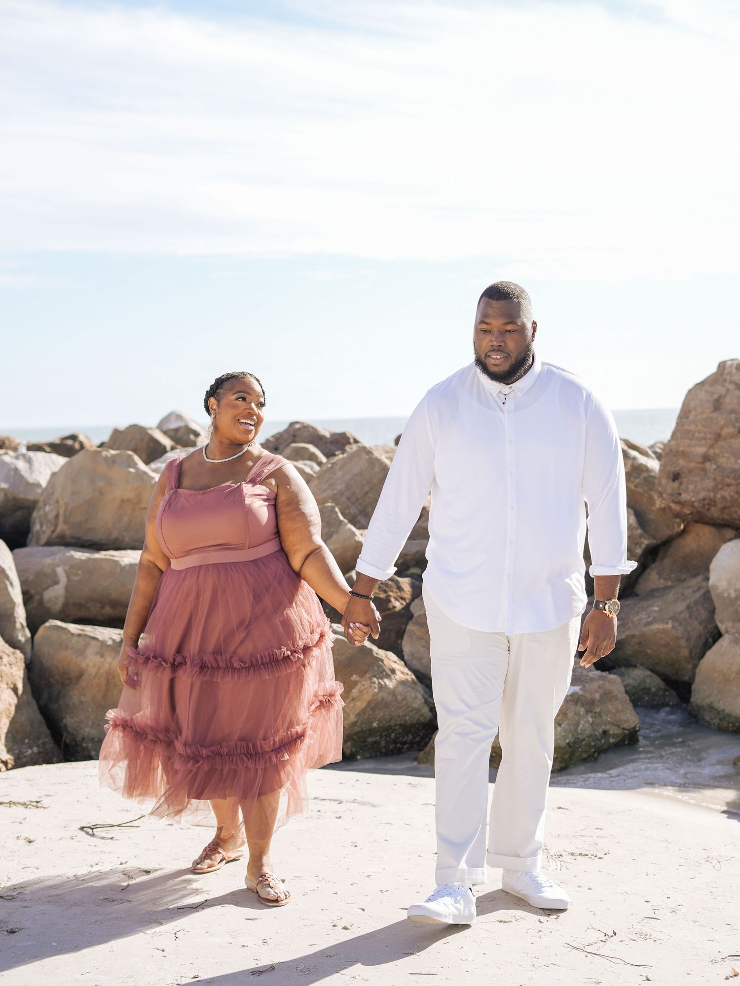 Couple in romantic beach photo, woman in pink tulle dress embracing man in white outfit by the ocean.