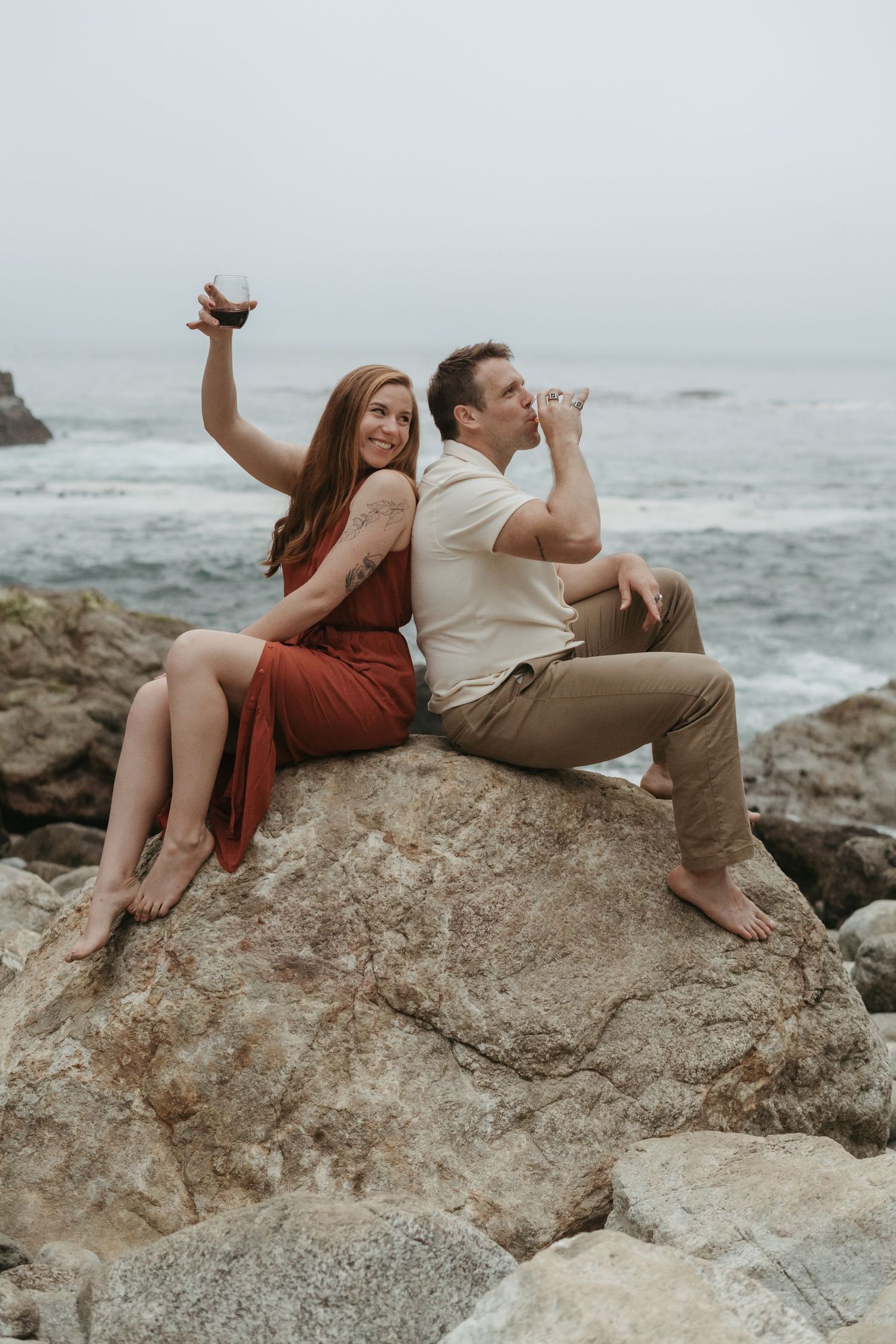 Couple sitting on wooden pallet by rocky ocean shore, toasting with ice cream cones beside sunflower bouquet.