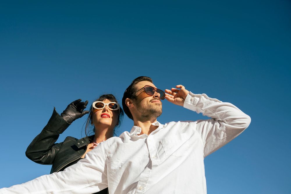 Stylish couple wearing sunglasses posing together against a bright blue sky.