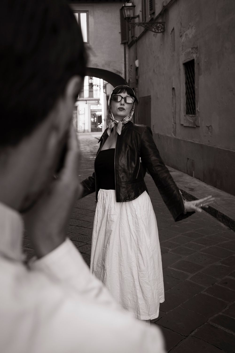 Black and white photo of a woman in leather jacket and white skirt on a cobblestone street.