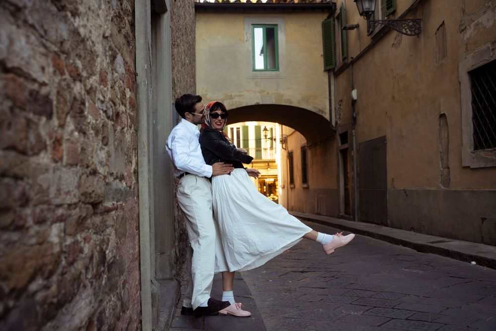 Couple embracing in a charming Italian alleyway, woman lifted off ground in white skirt.