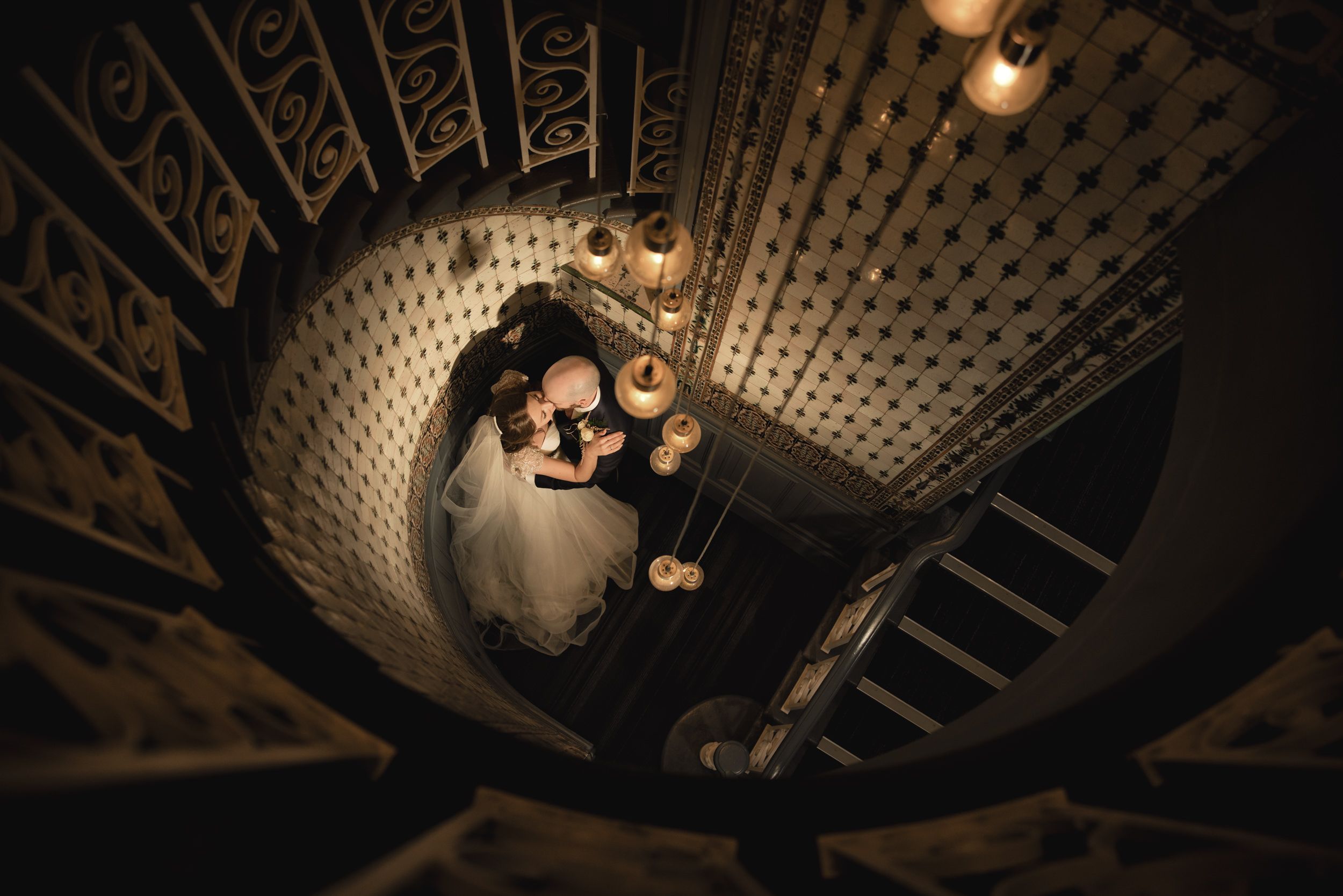Aerial view of bride and groom embracing on ornate spiral staircase with warm glowing lights.