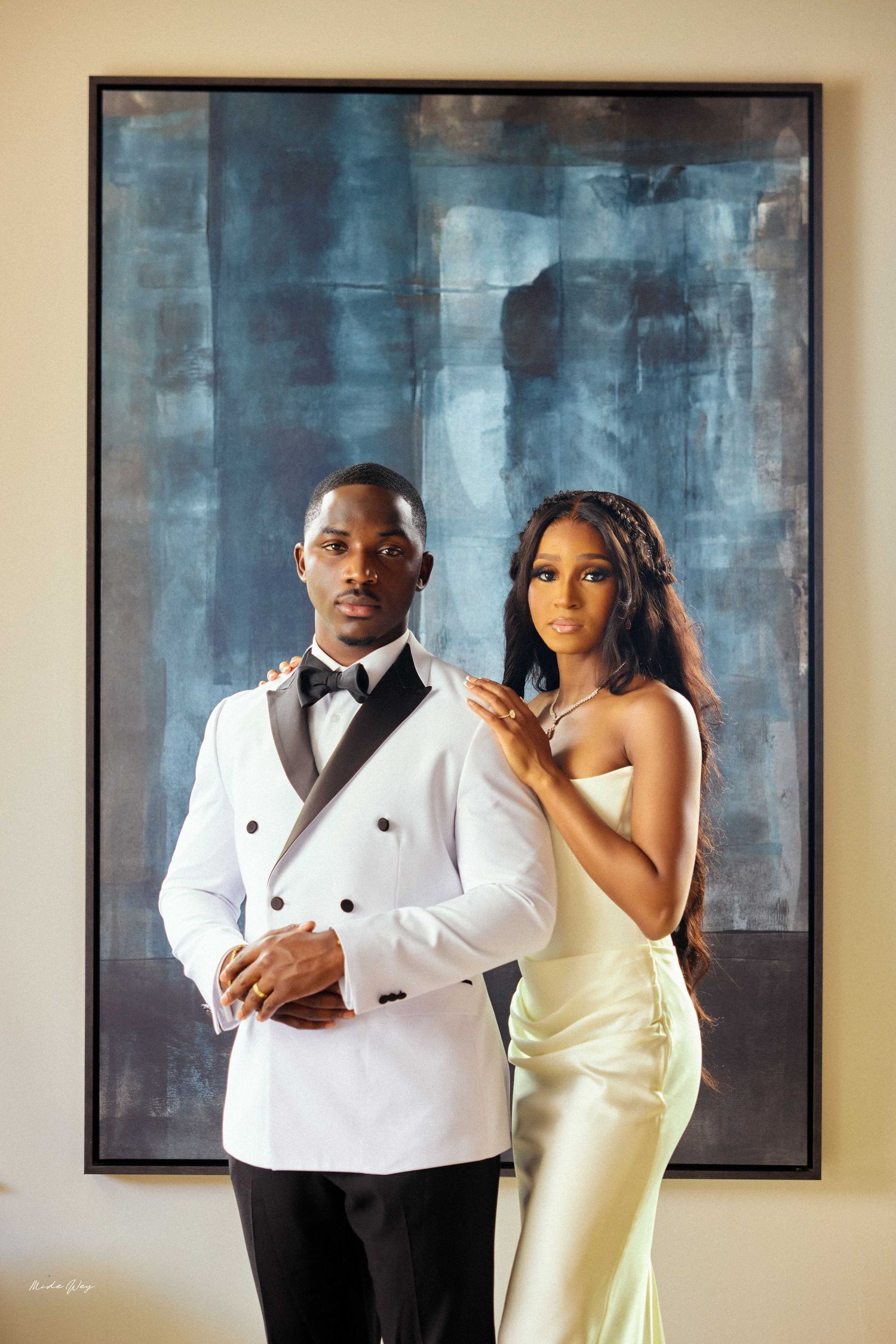 Elegant couple posing before abstract blue painting, man in white tuxedo, woman in gold satin gown.