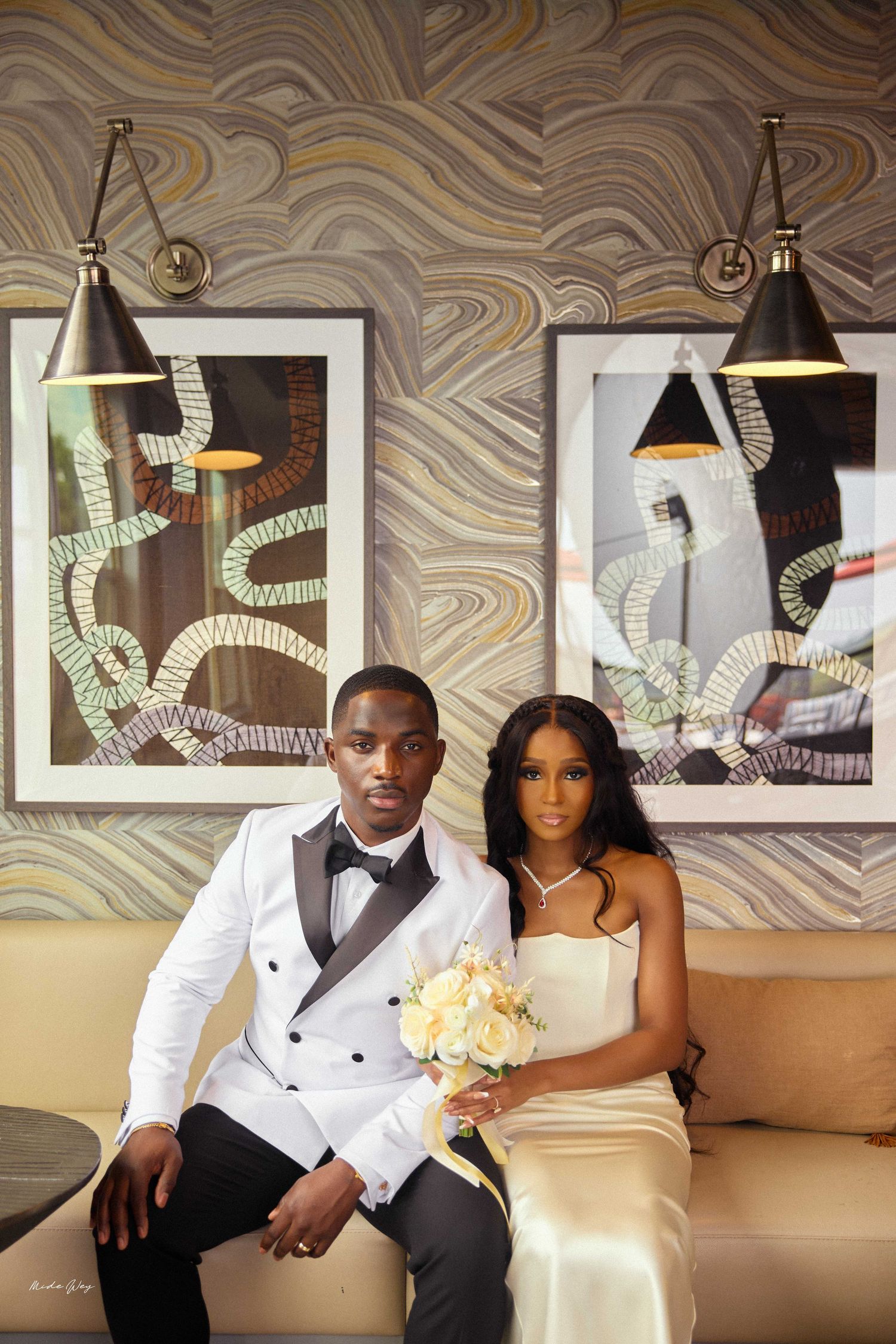 Elegant couple in wedding attire sitting on a sofa, groom in white tuxedo, bride holding bouquet.