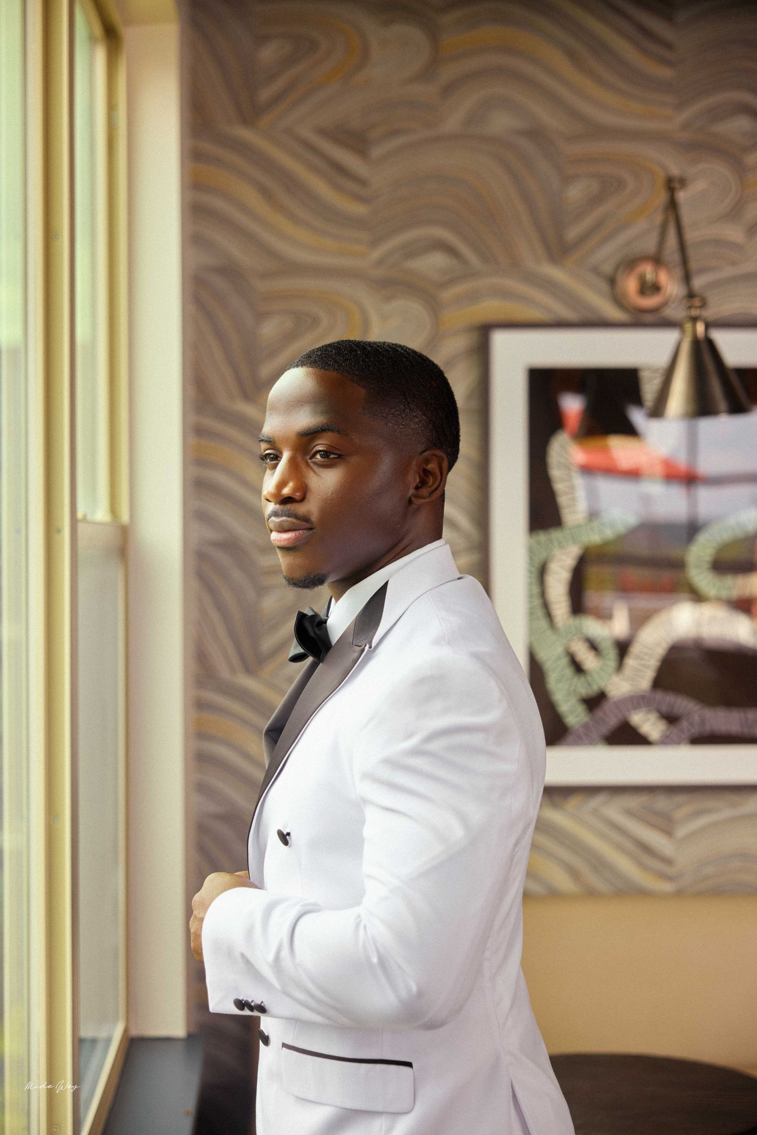 A young Black man in a white tuxedo with black bow tie stands by a window in an elegant room.