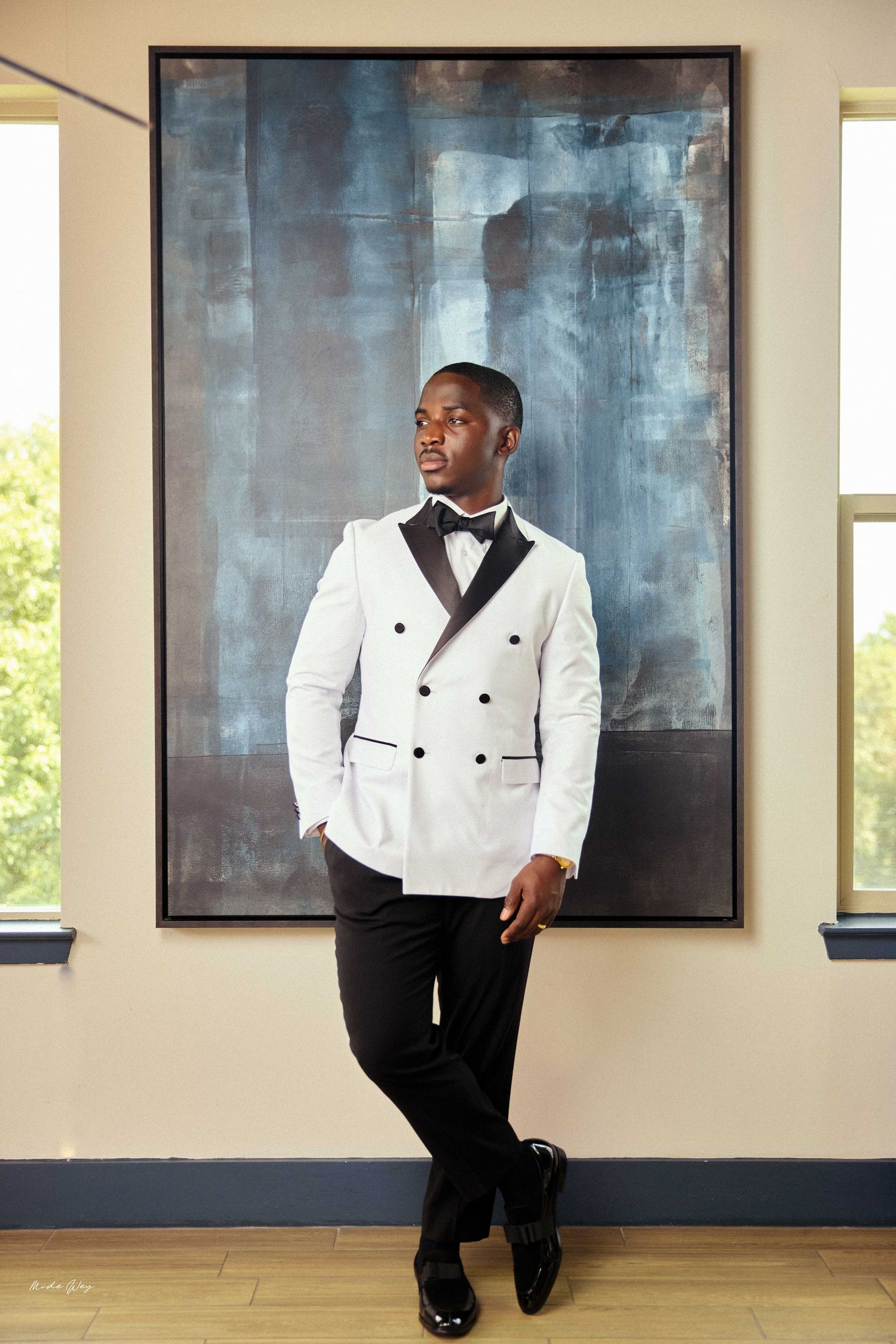 Man in white double-breasted tuxedo jacket with bow tie poses confidently before large abstract painting.