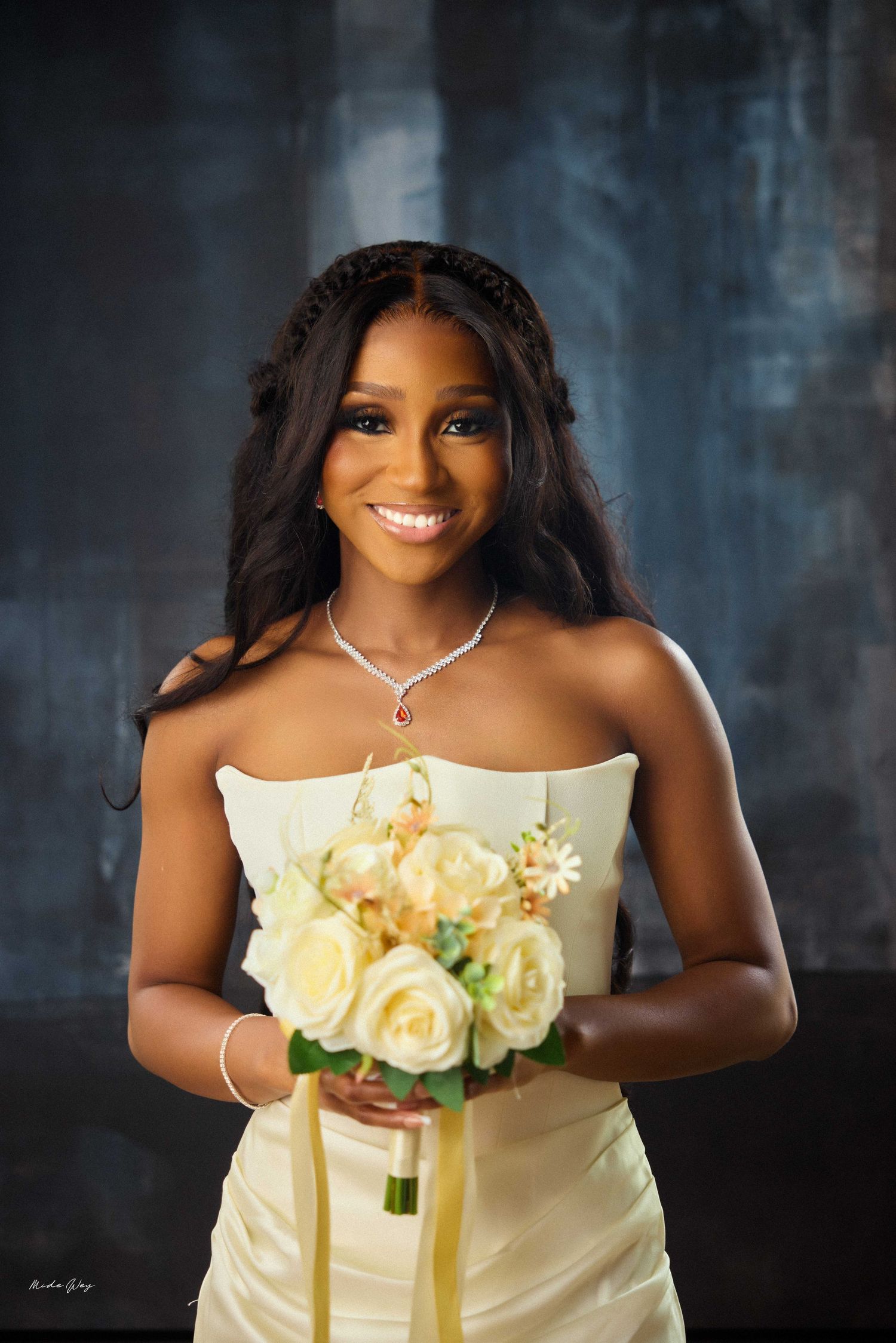 Smiling bride in strapless ivory gown holding white rose bouquet, wearing diamond necklace, posing against dark backdrop.