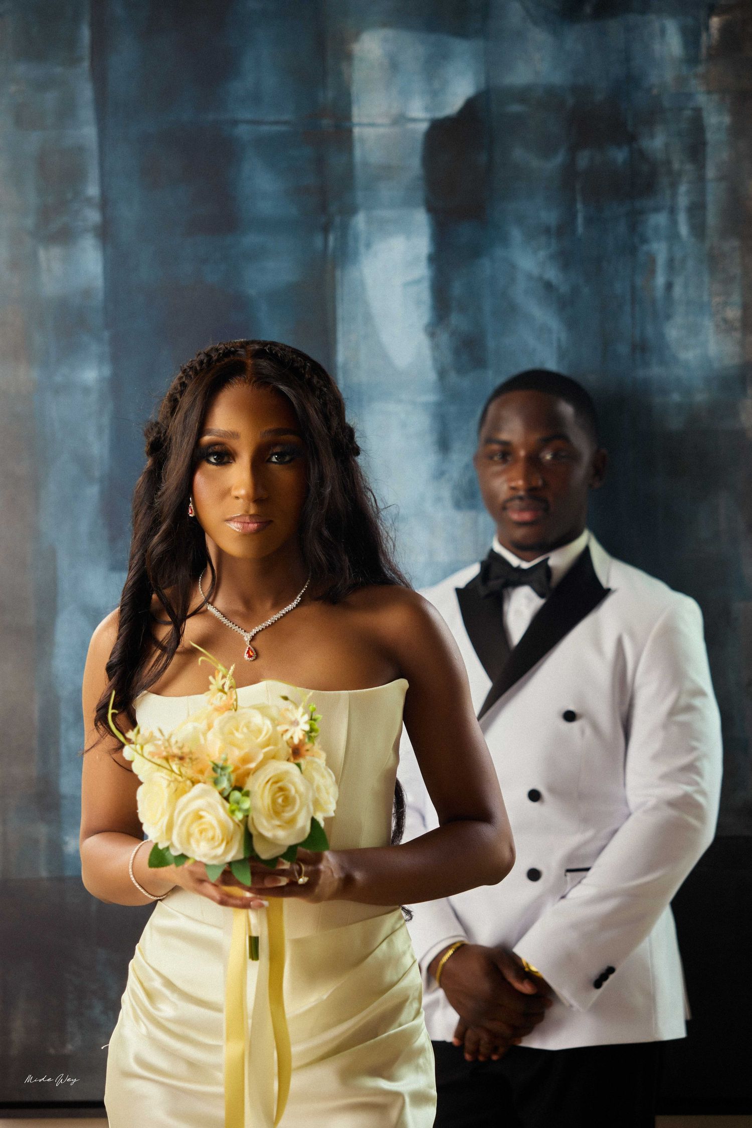 Bride in strapless ivory gown holding yellow roses bouquet with groom in white tuxedo posing before dark blue backdrop.