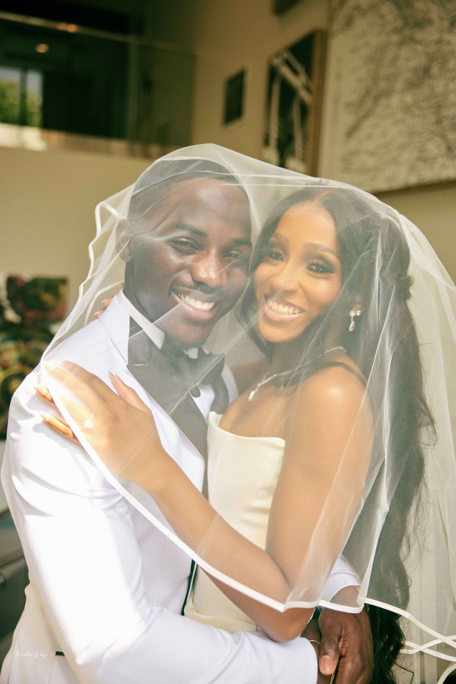 Happy newlywed couple smiling under a bridal veil, groom in white suit and bride in strapless wedding dress.