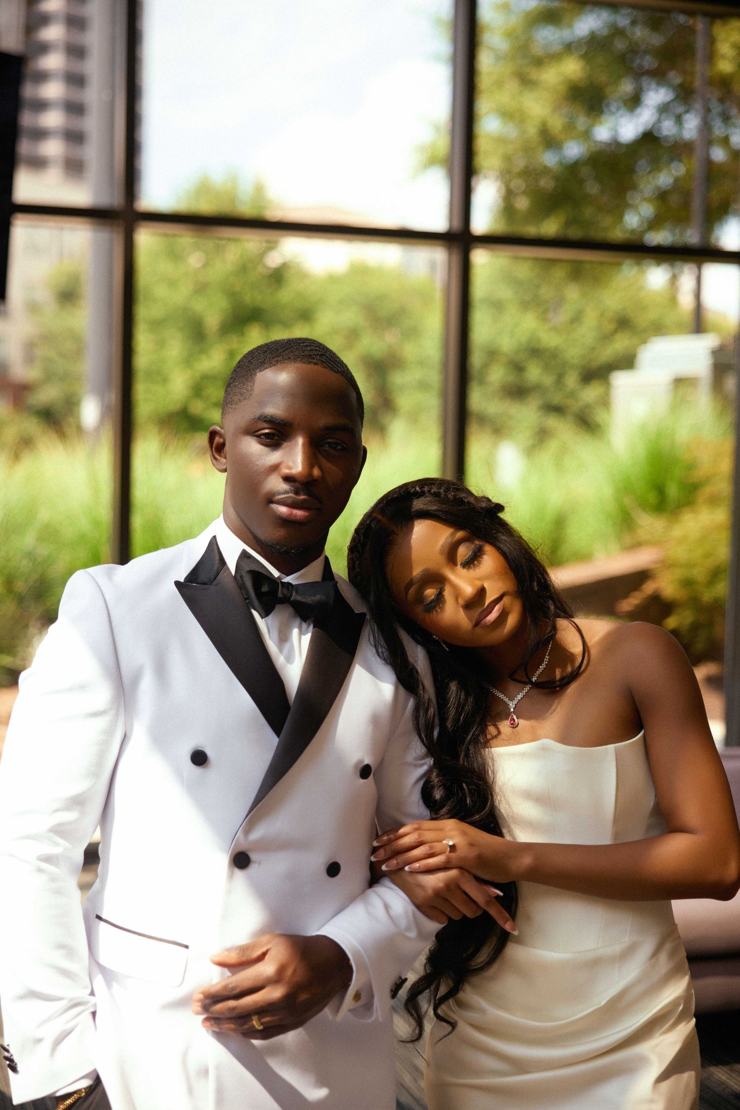 Elegant Black couple in wedding attire, groom in white tuxedo, bride in satin dress, posing together indoors.