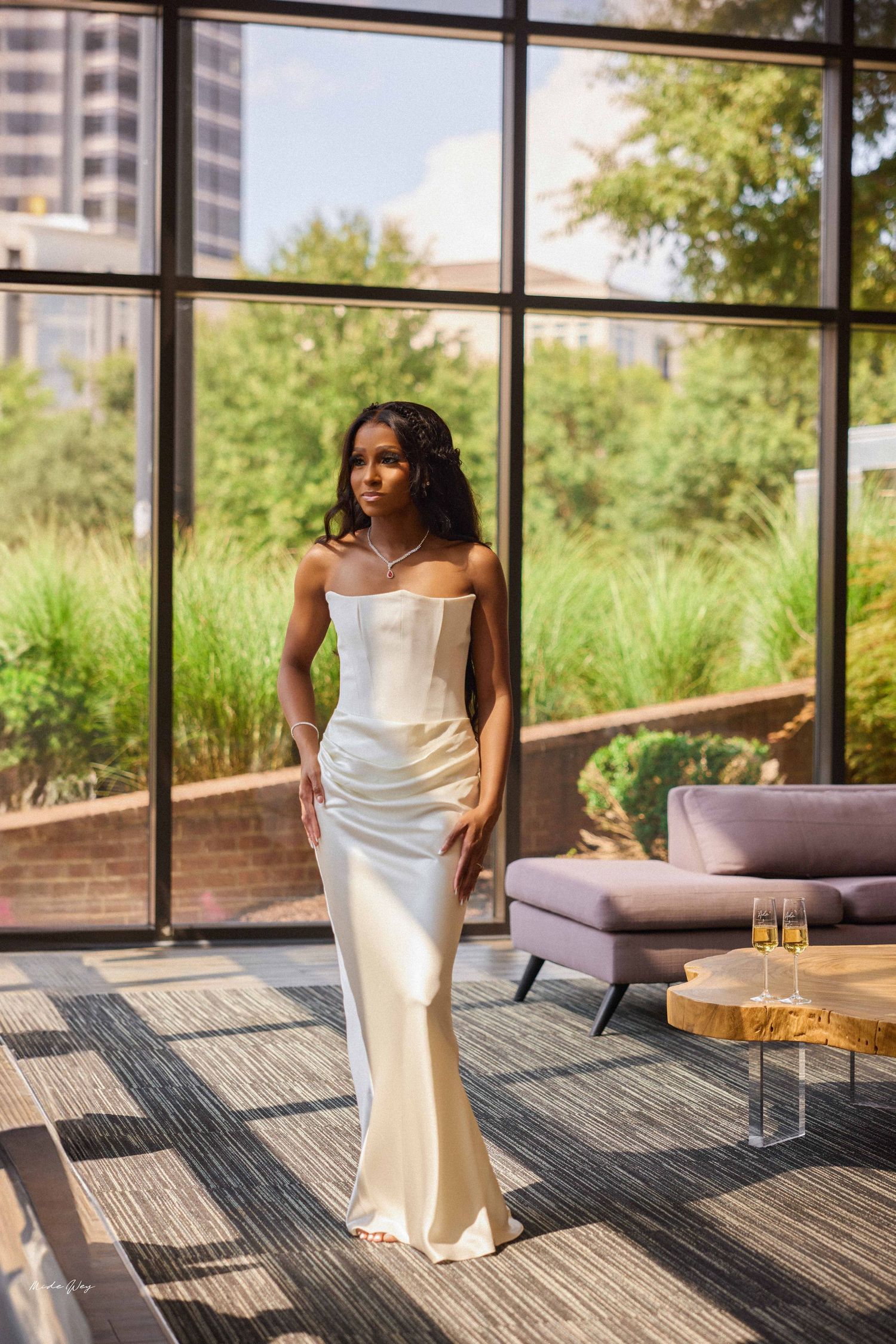 Elegant bride in strapless ivory satin gown poses near floor-to-ceiling windows with lush greenery backdrop.