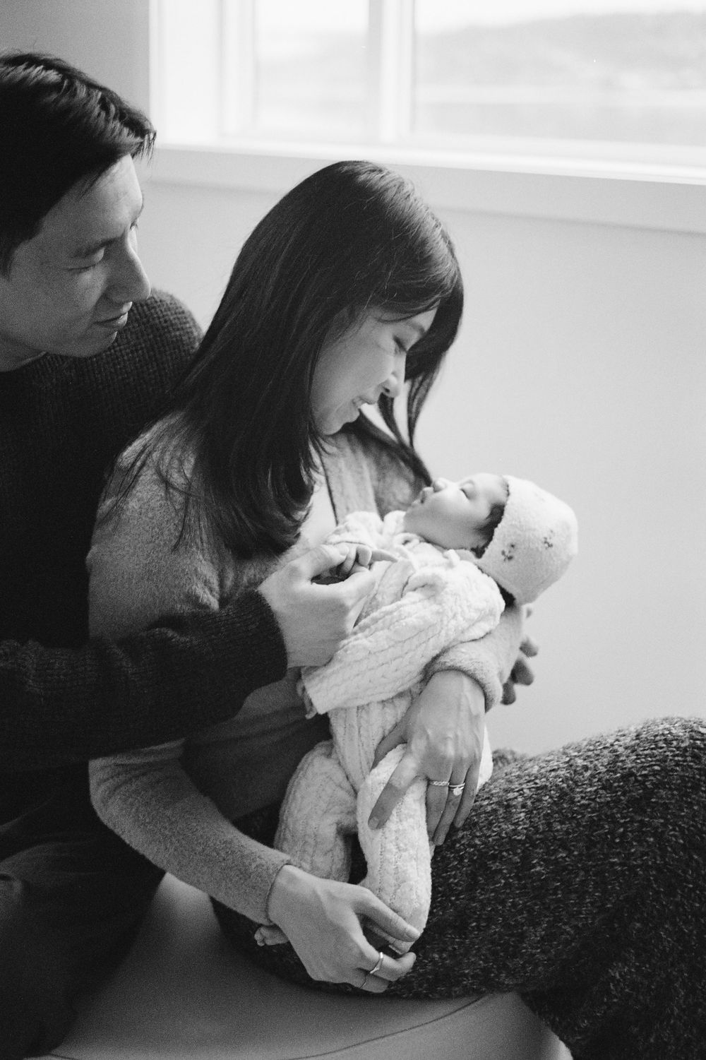 Black and white photo of new parents lovingly gazing at their swaddled newborn baby indoors.