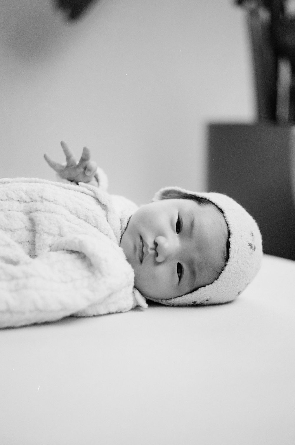 Black and white photo of a newborn baby lying down wearing a soft hat and cozy outfit, one hand raised.