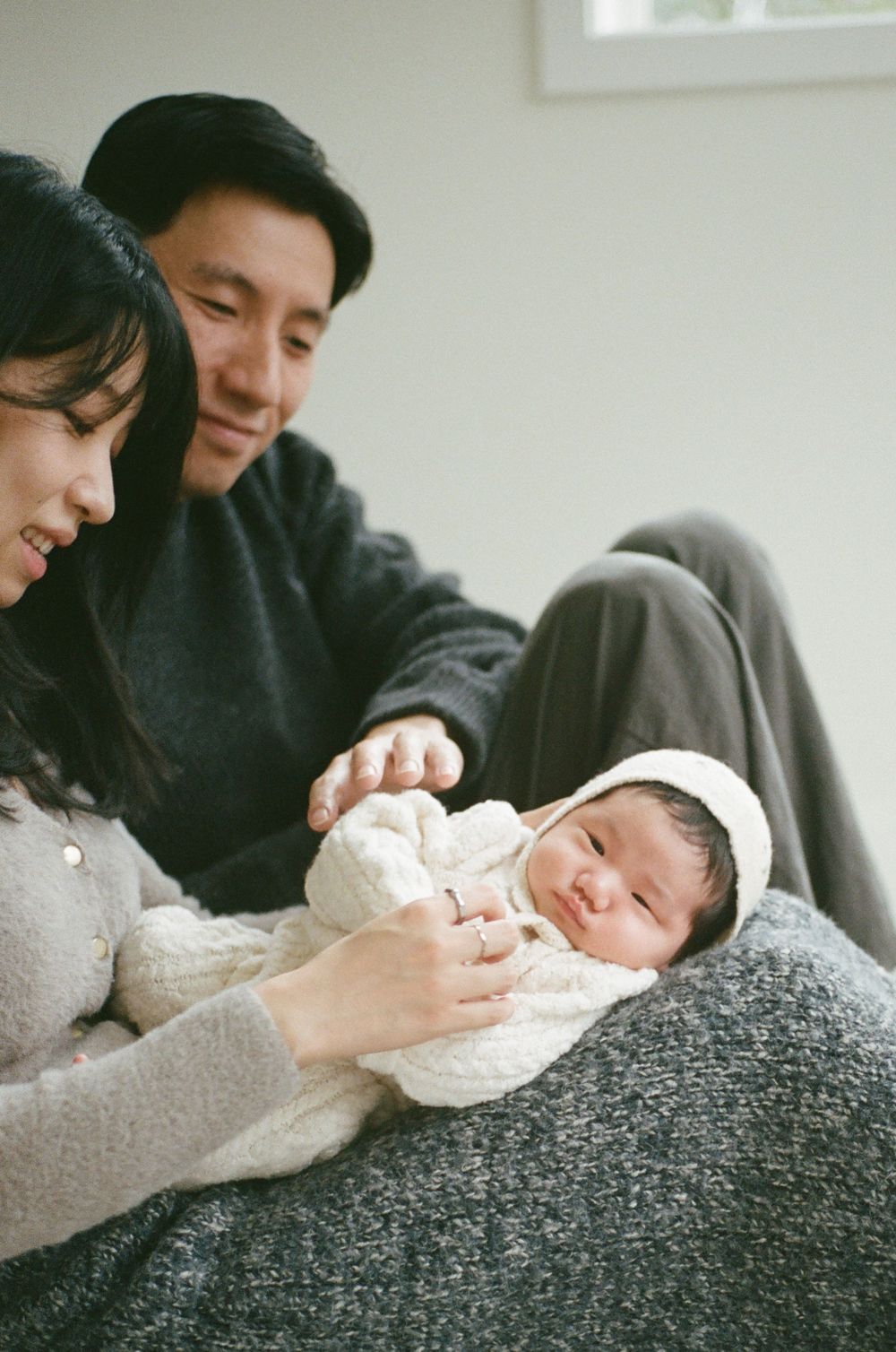 New parents sitting on floor cradling their swaddled newborn baby wrapped in white blanket with hood.