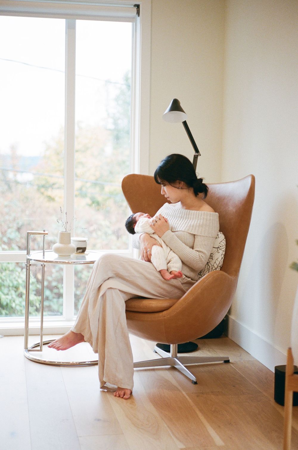 A mother sits in a brown leather egg chair, cradling her newborn baby in a bright, modern living room.