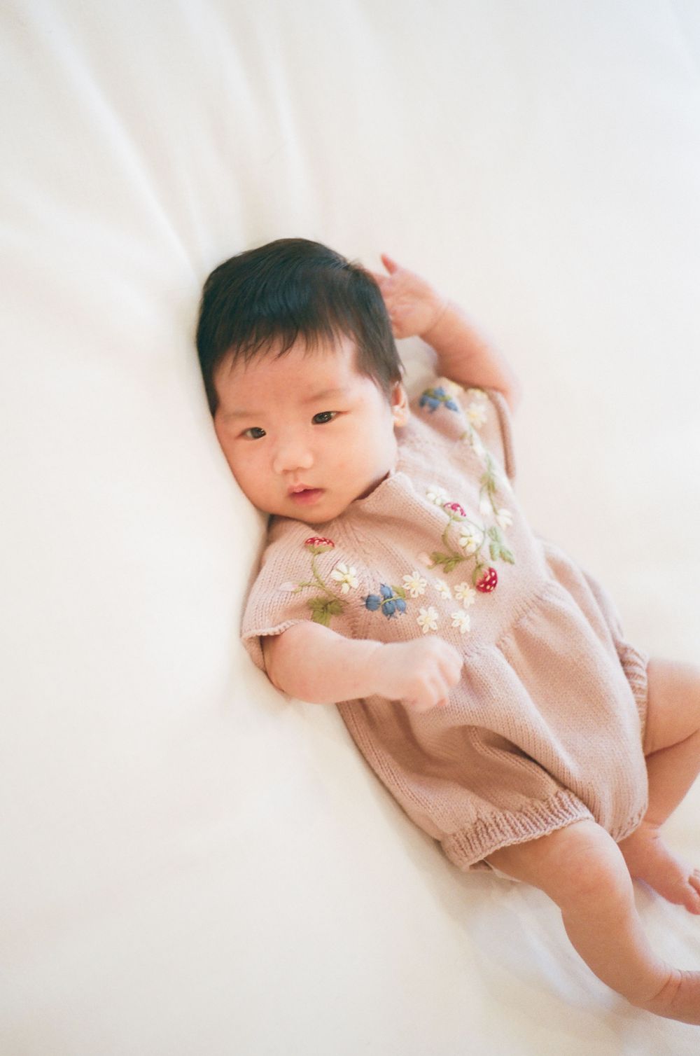 A cute Asian baby lying on a white blanket wearing a pink floral embroidered romper.