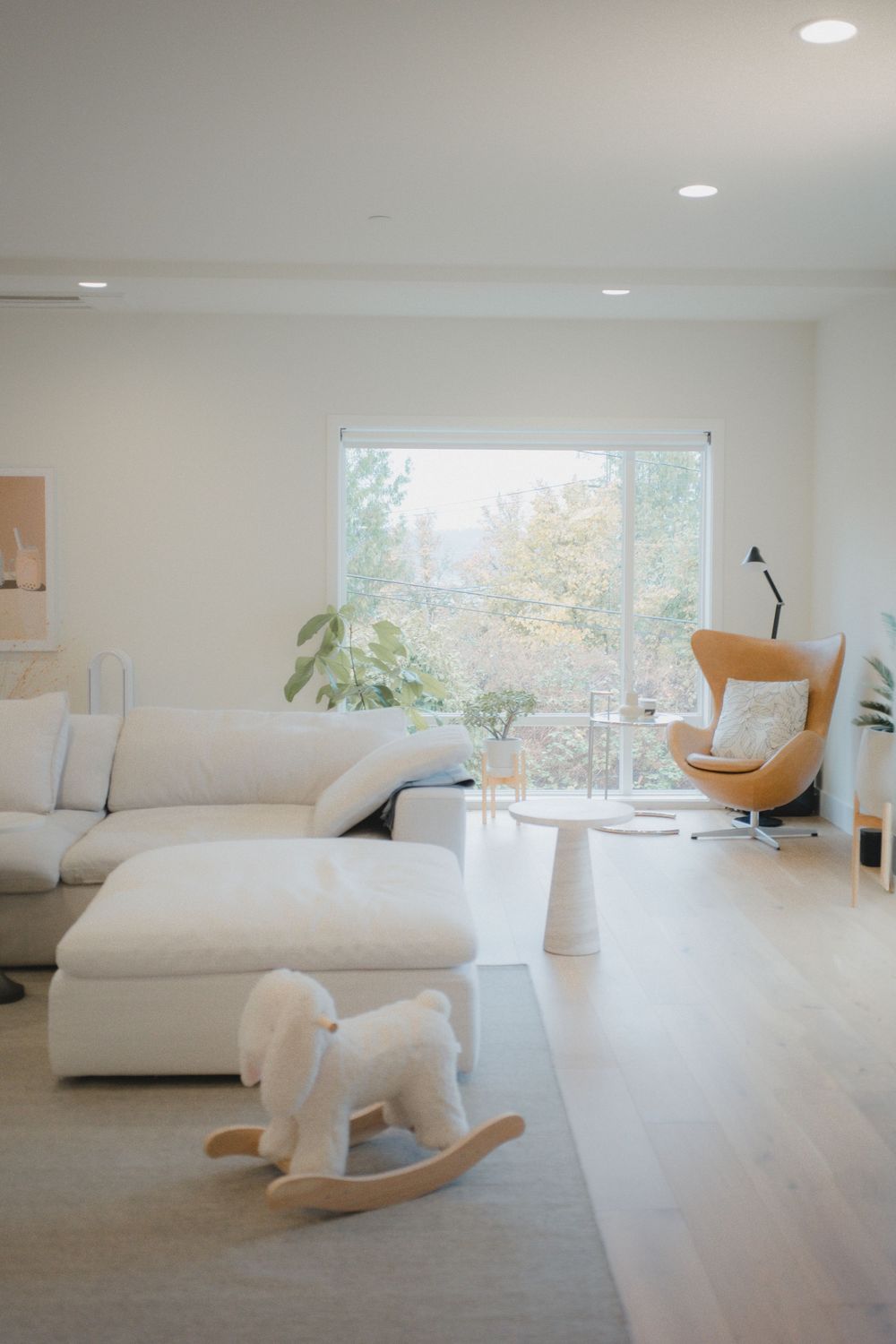 Minimalist white living room with plush sofa, rocking horse toy, egg chair, and large window overlooking green trees.