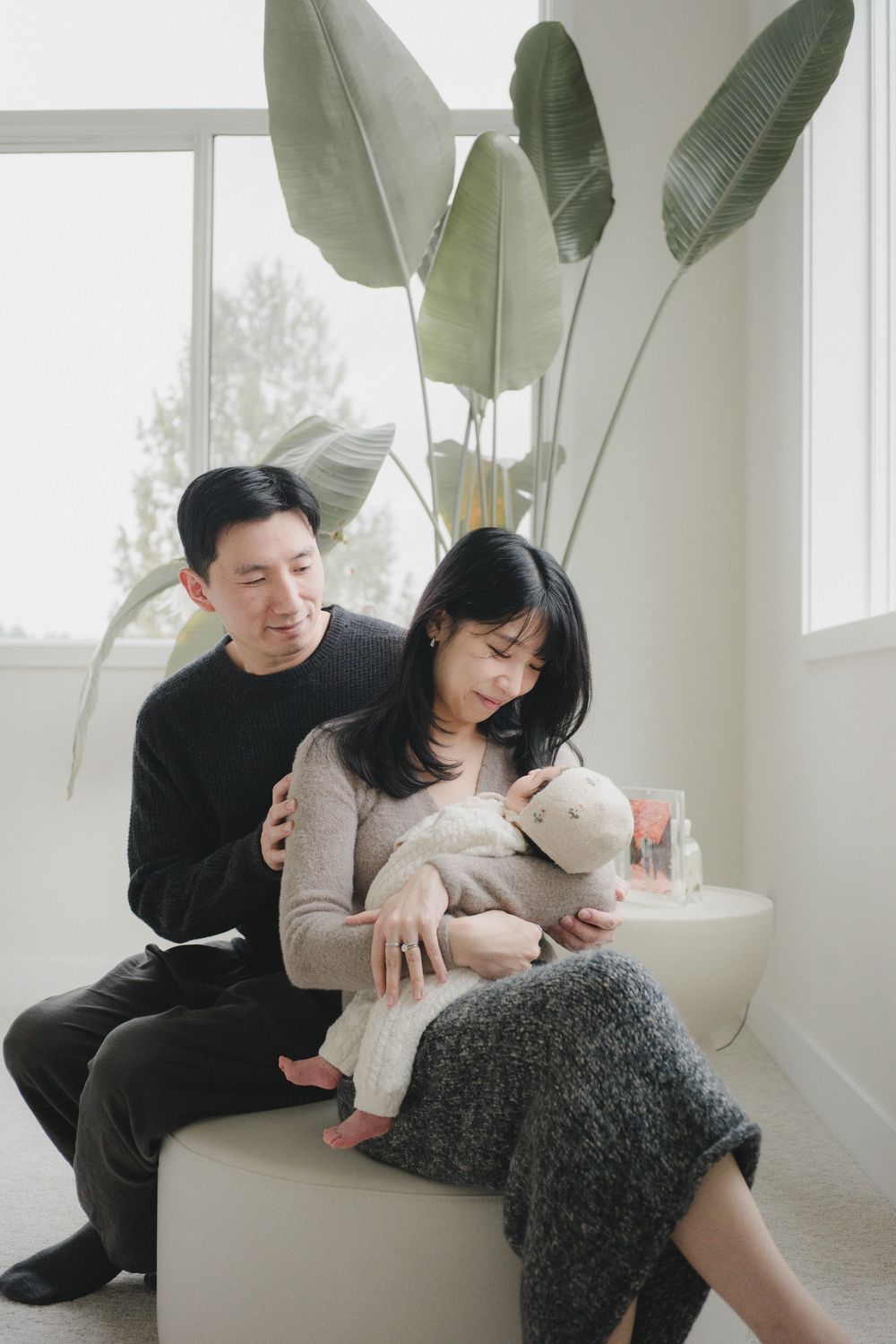A couple sits by bright windows with a large tropical plant, lovingly cradling their newborn baby with a stuffed animal.