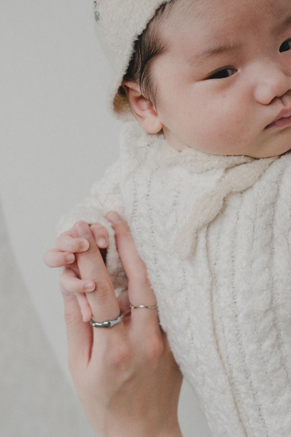 Newborn baby in cream knit wrap grasping adult finger wearing silver rings in soft natural light.