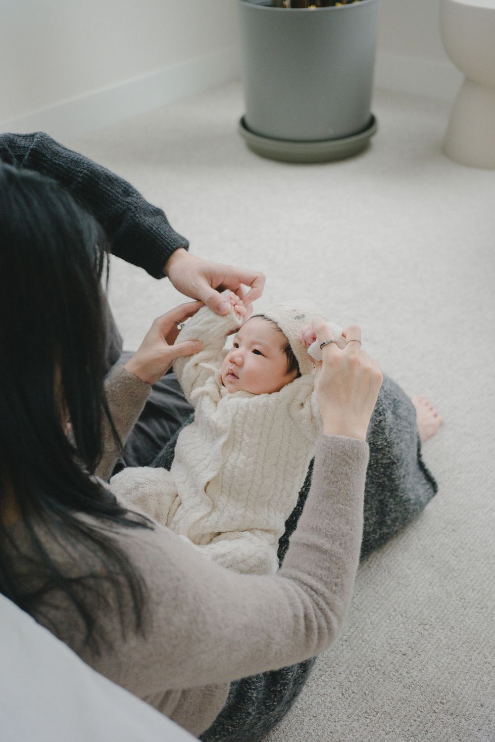 A mother tenderly holds her newborn baby in a white knit outfit while sitting on a soft carpet floor.