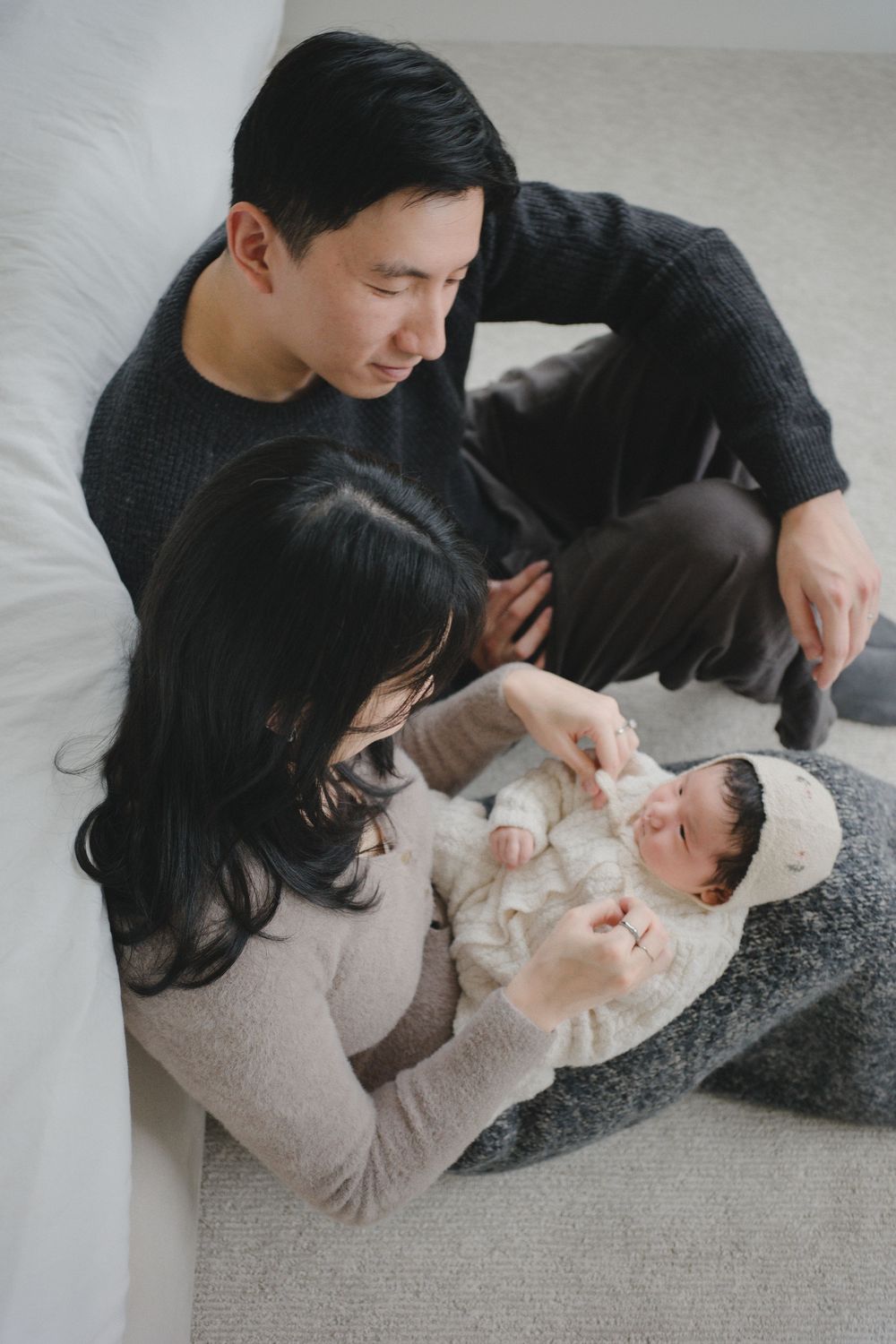 Asian couple sitting on floor with newborn baby in white outfit, sharing a tender family moment together.