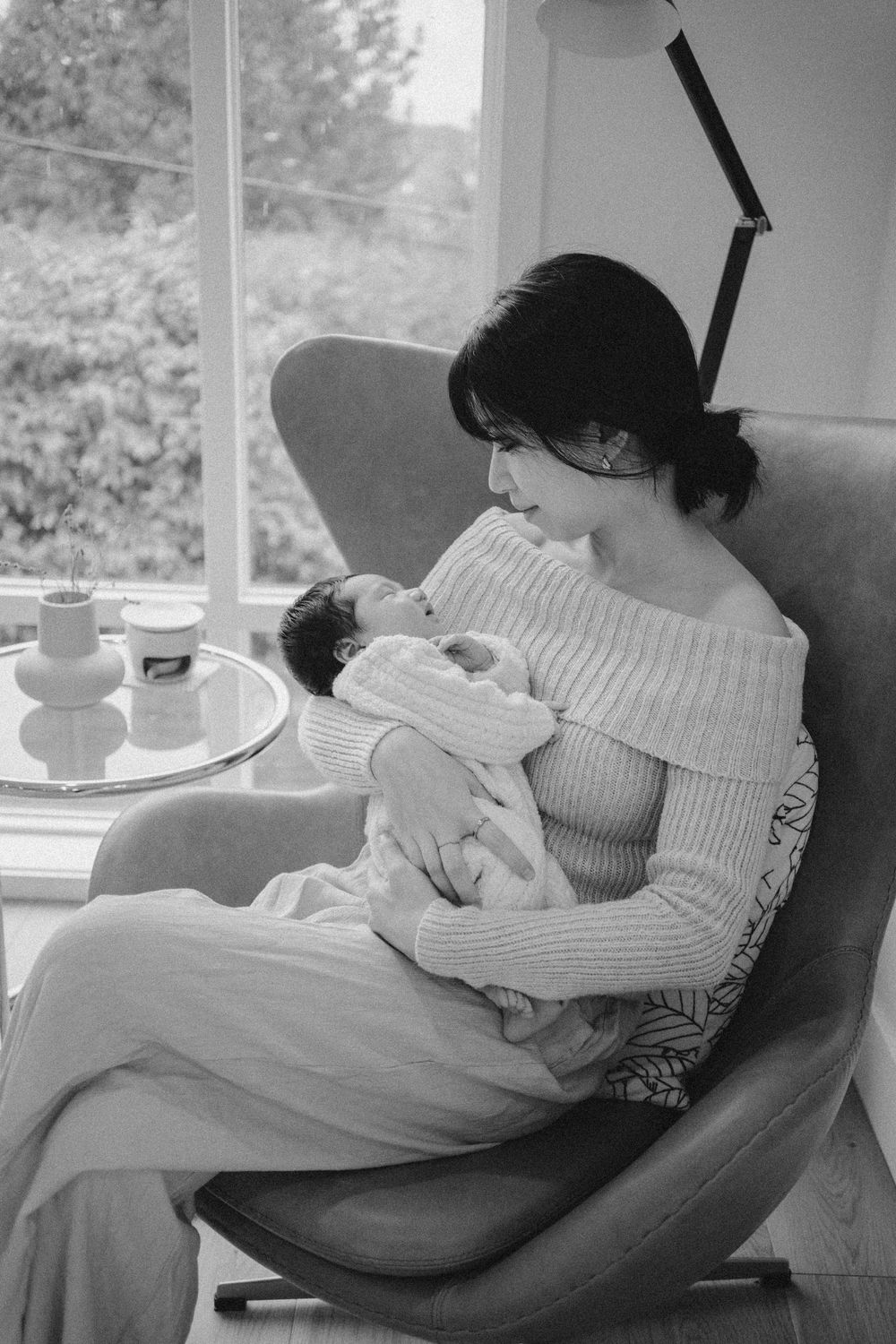 Black and white photo of a mother cradling her newborn baby in an armchair by a window.