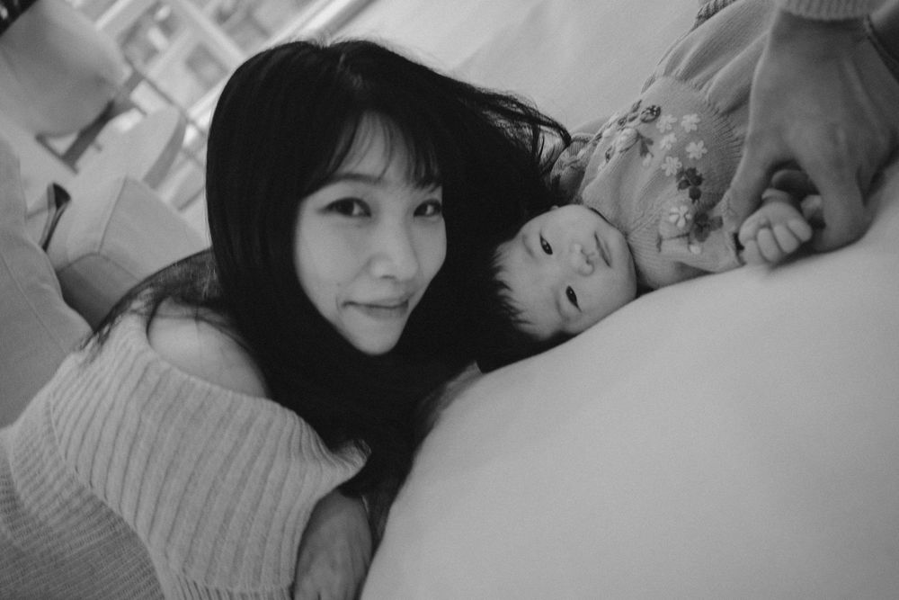 Black and white photo of a young Asian woman smiling beside a sleeping baby on a soft surface.