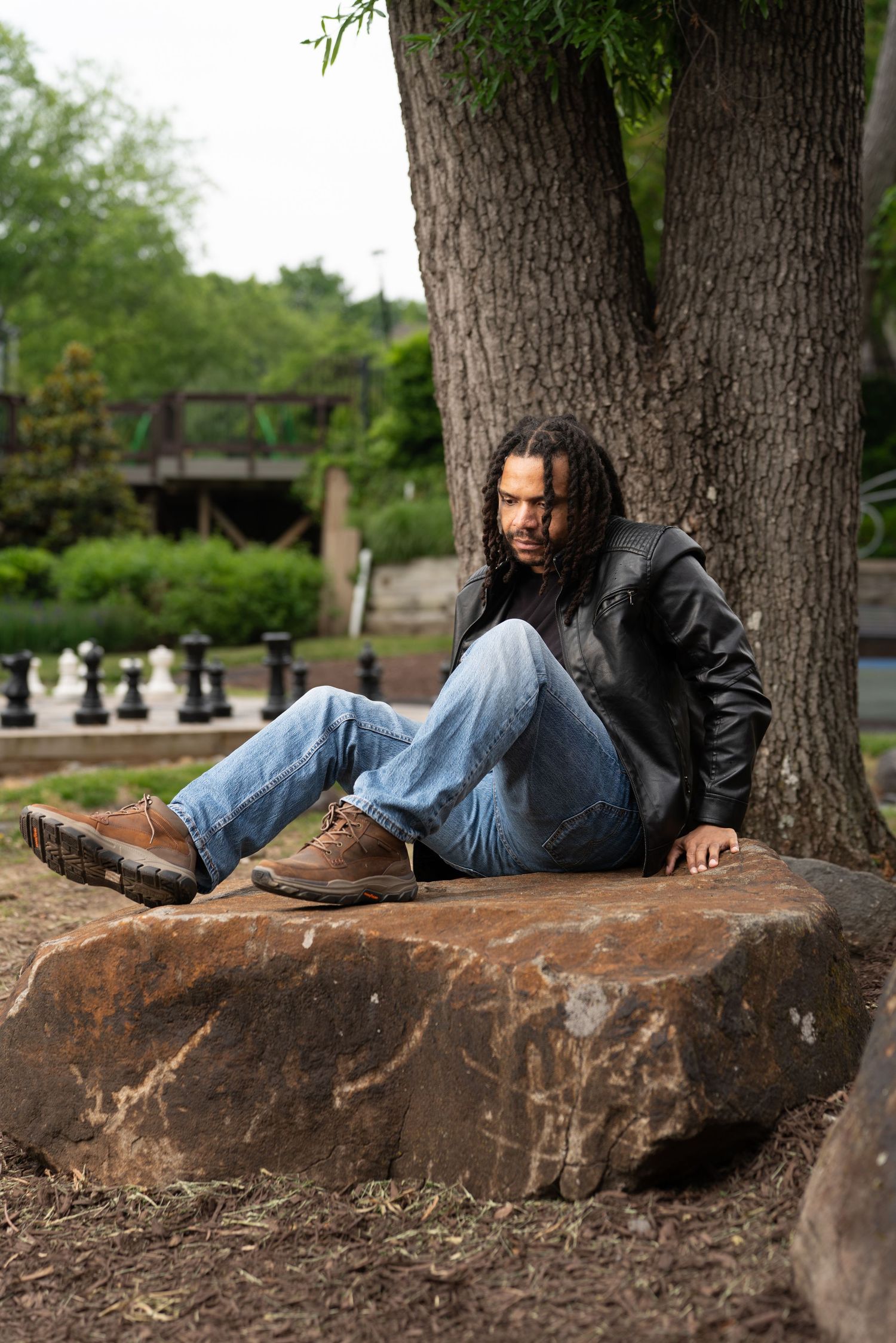 A man with dreadlocks in a leather jacket and jeans sits on a large rock beneath a tree in a park.