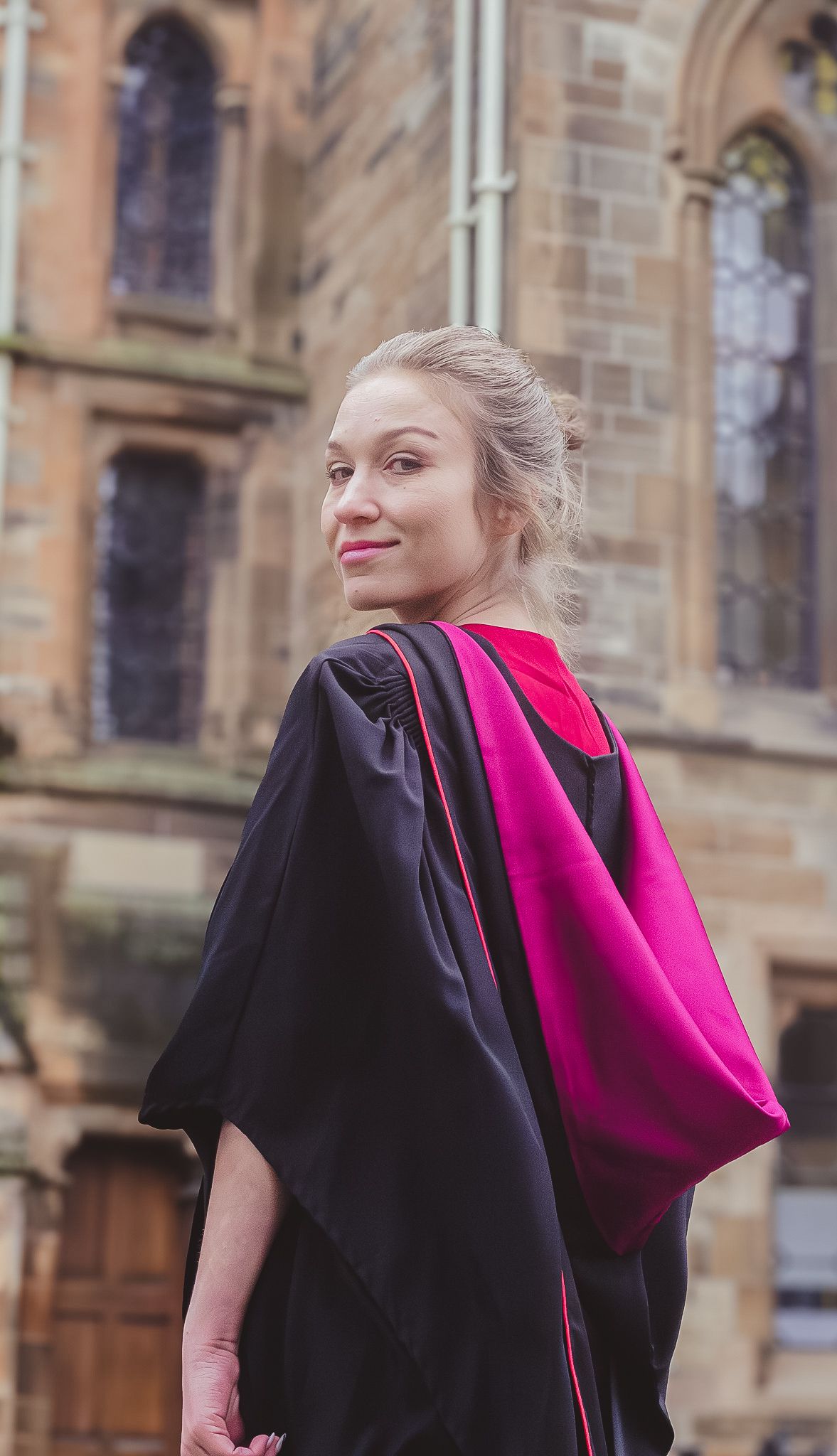 A smiling woman in black and pink graduation robes stands before a historic stone university building.