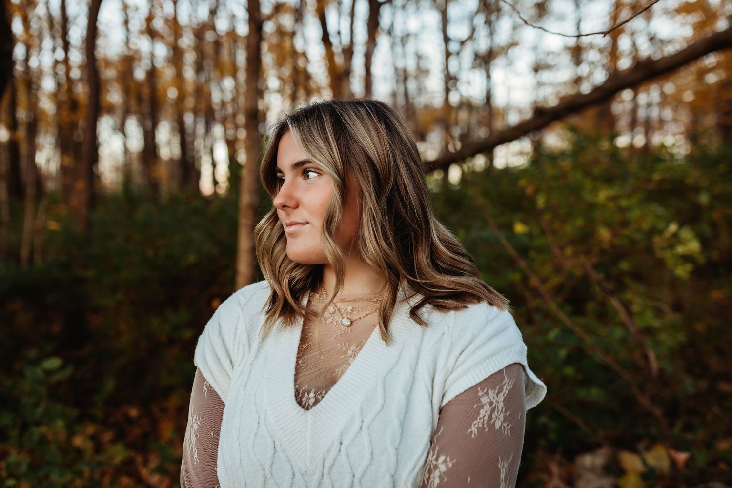 A person in a white lace dress poses outdoors among autumn trees during a golden hour photoshoot.