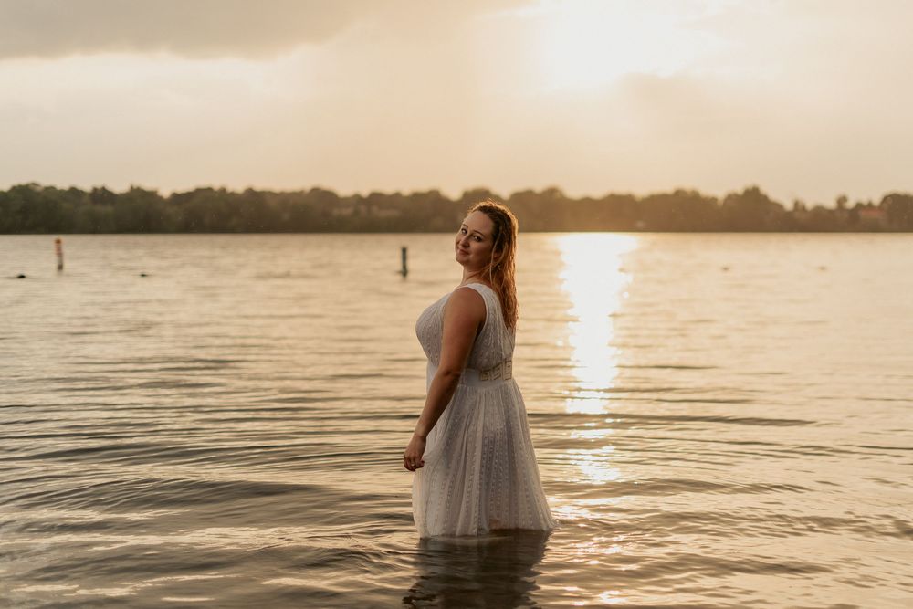Figure in a white dress stands in calm water at sunset creating a dreamy silhouette against the golden light.