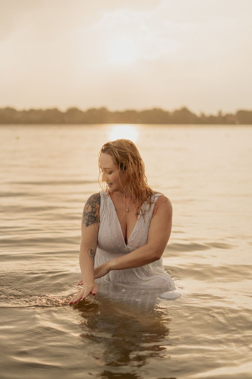 A person in a gray dress wades through tranquil waters at sunset creating ripples in the golden light.
