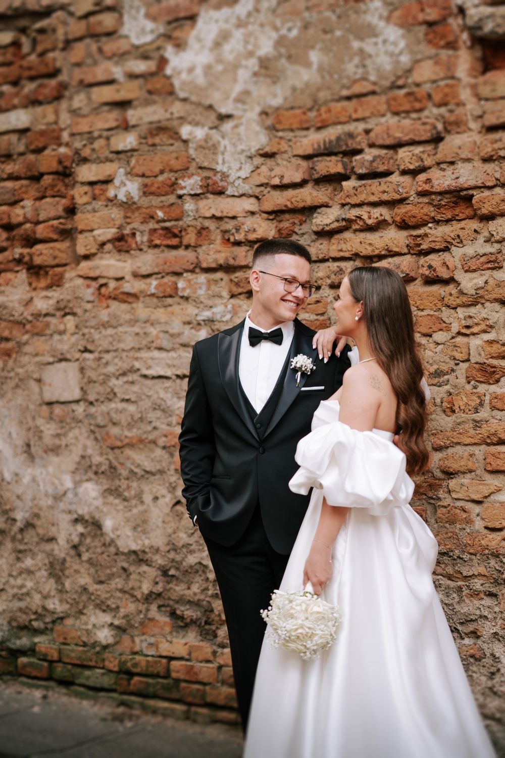 Newlyweds pose against a rustic brick wall during their wedding photoshoot.