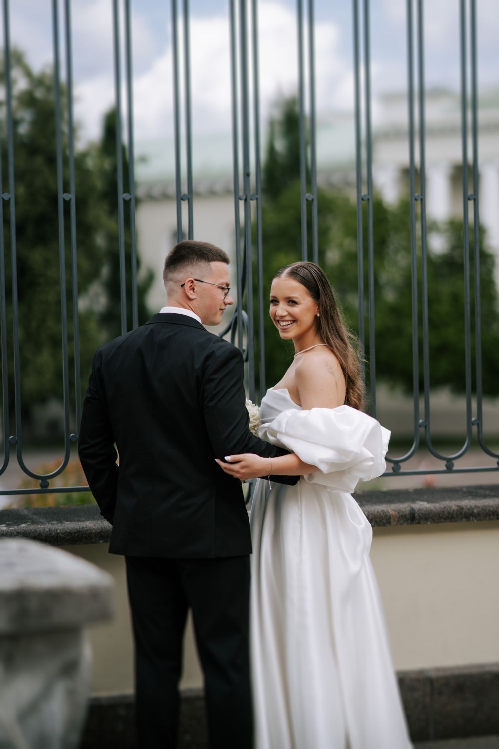 Wedding couple poses playfully by metal railings during an outdoor photoshoot.