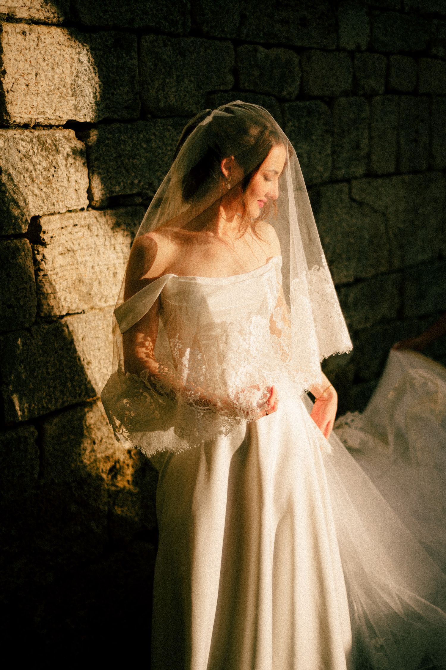A bride in a flowing white lace wedding dress and veil stands against a dark stone wall in dramatic lighting.