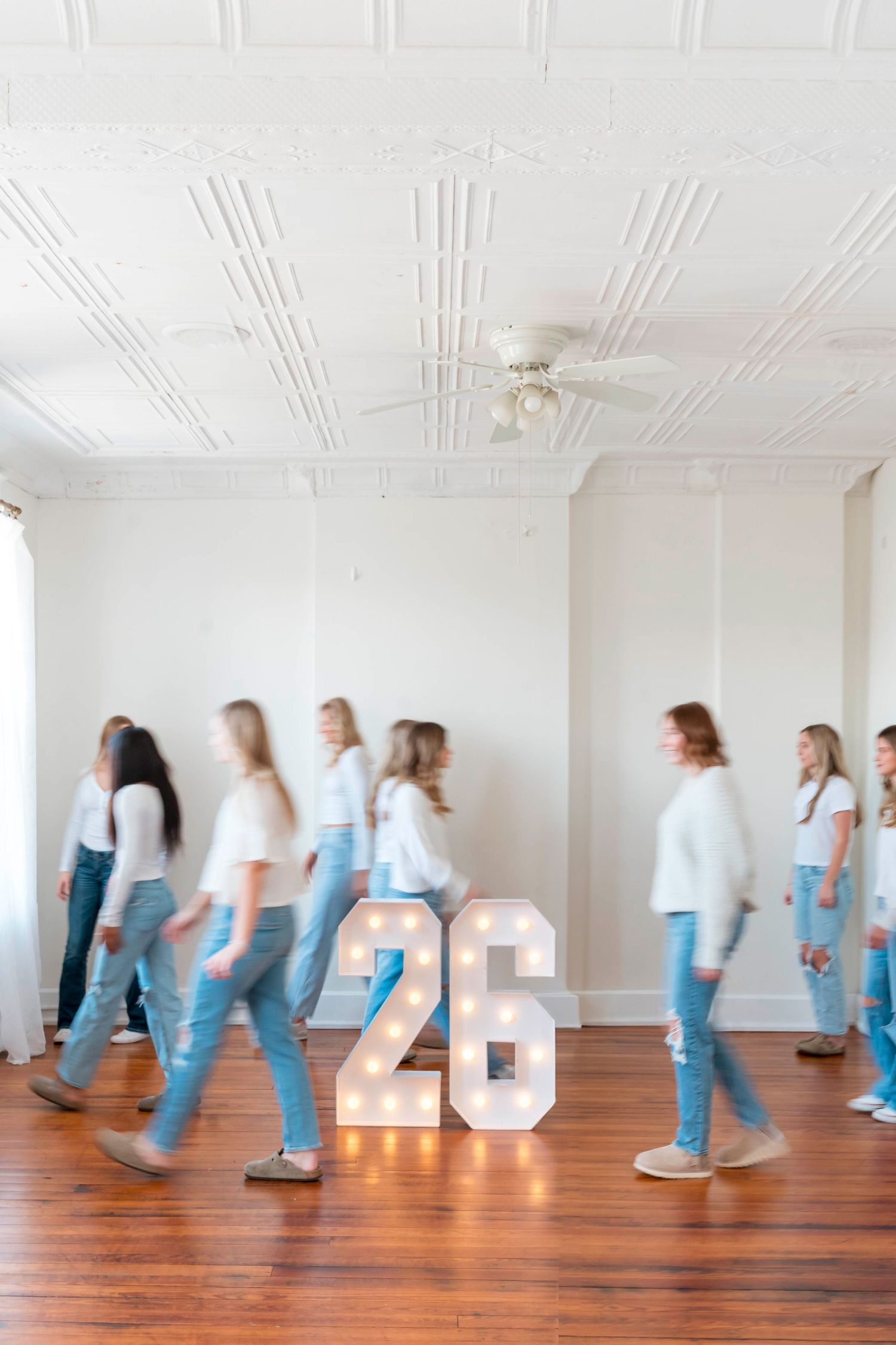 Motion blur of people walking past large white numbers on hardwood floor in bright minimalist gallery space.