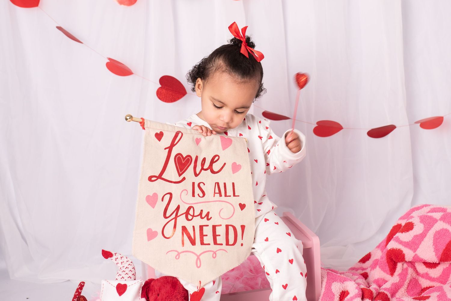 Pink and red heart-shaped confetti falls around a small child in a white dress during a Valentine's Day photo shoot.