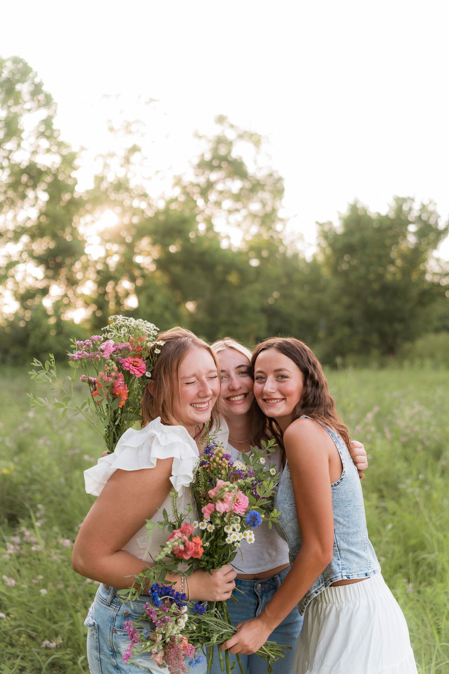 Three people embrace while laughing together in a sunlit meadow with wildflowers and greenery.