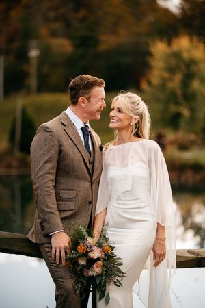 A romantic wedding couple shares tender moments in autumn by a lake wearing a white dress and brown tweed suit.