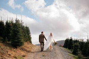 Two people walk along a scenic mountain path with evergreen trees and dramatic cloudy skies in the background.