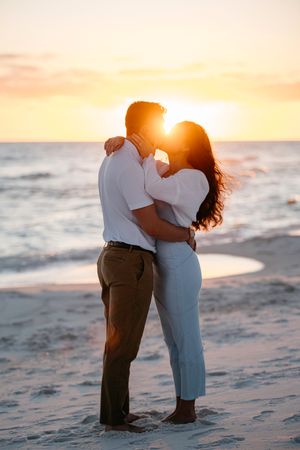 A romantic couple embraces on a sandy beach at sunset with golden sunlight streaming between them.