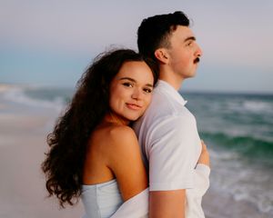 A couple embraces on a beach at sunset with waves crashing behind them.