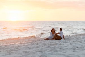 A couple sits together on the beach sand watching a beautiful orange sunset over the ocean.
