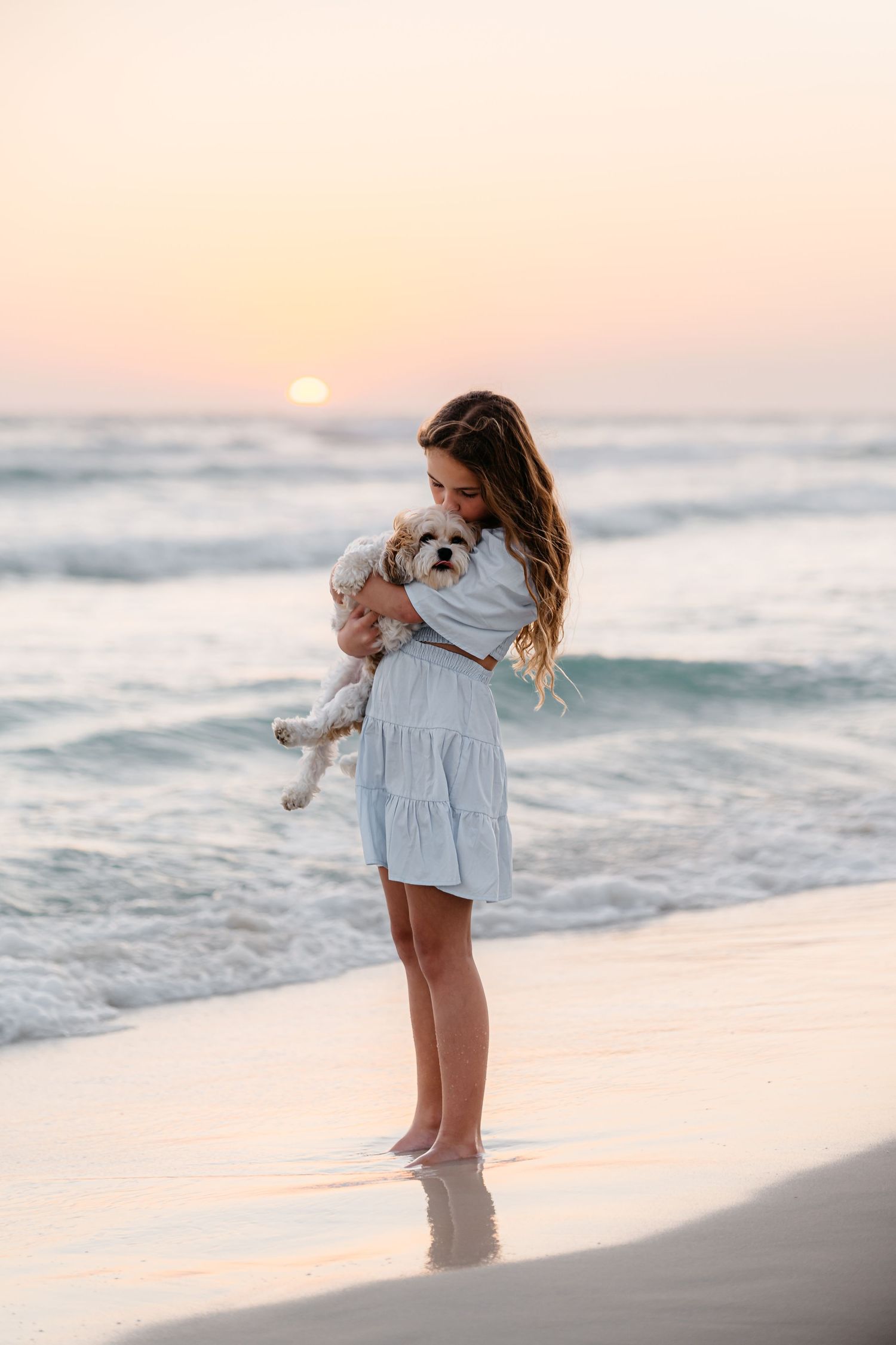 A person in a white dress holds a small fluffy dog while standing on a beach at sunset with ocean waves in the background.