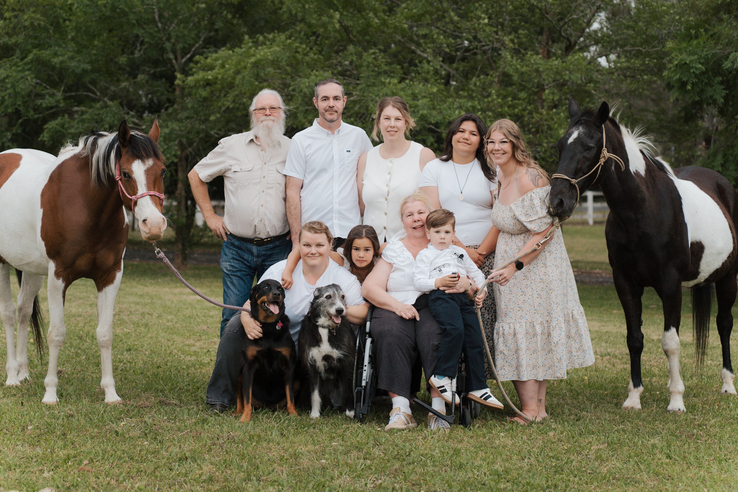 A group gathers in a grassy field with horses, wearing white and light-colored clothing for a rustic outdoor photoshoot in Gables Box Hill