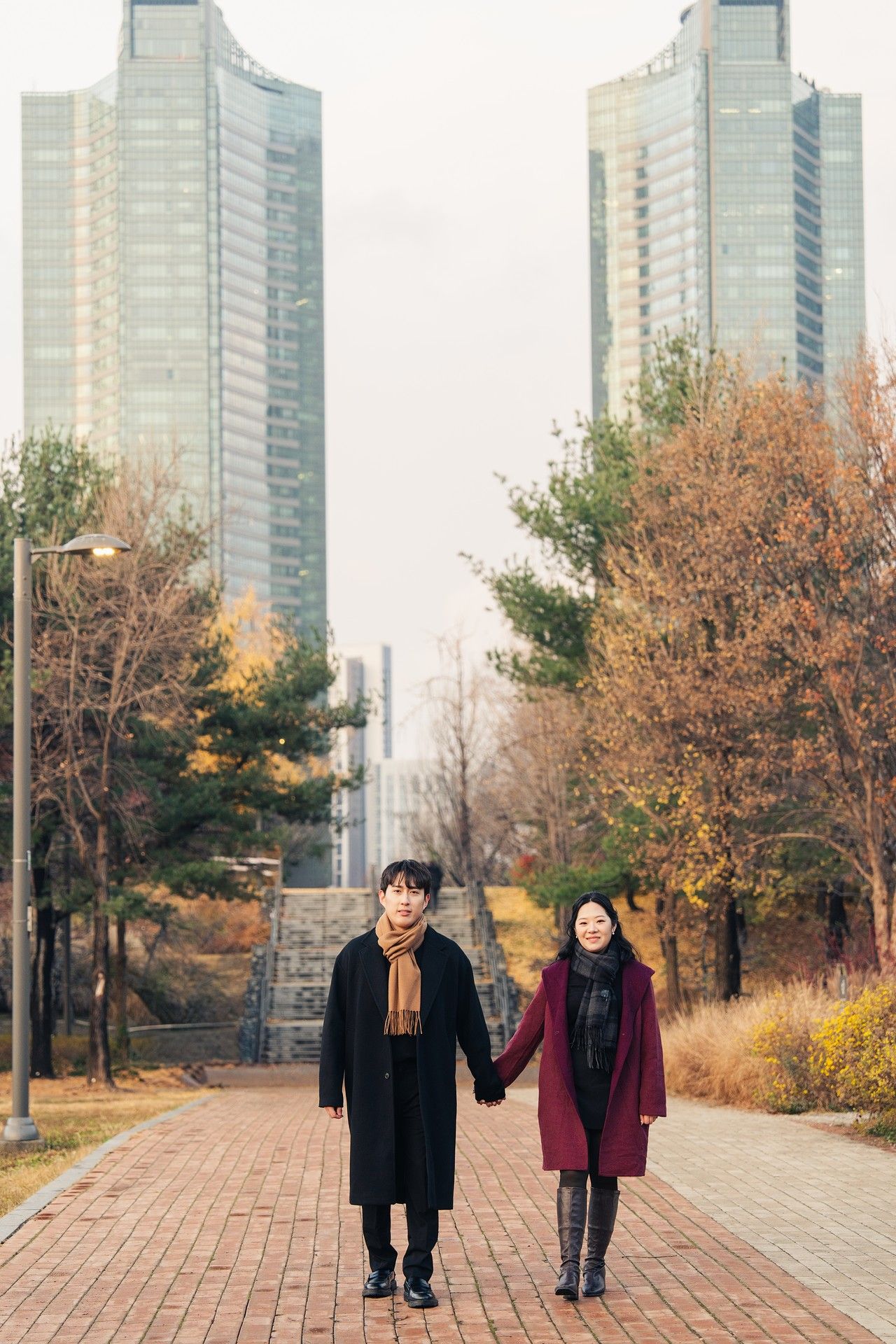 Two people walk together on a paved path surrounded by autumn trees and tall city buildings in the background.