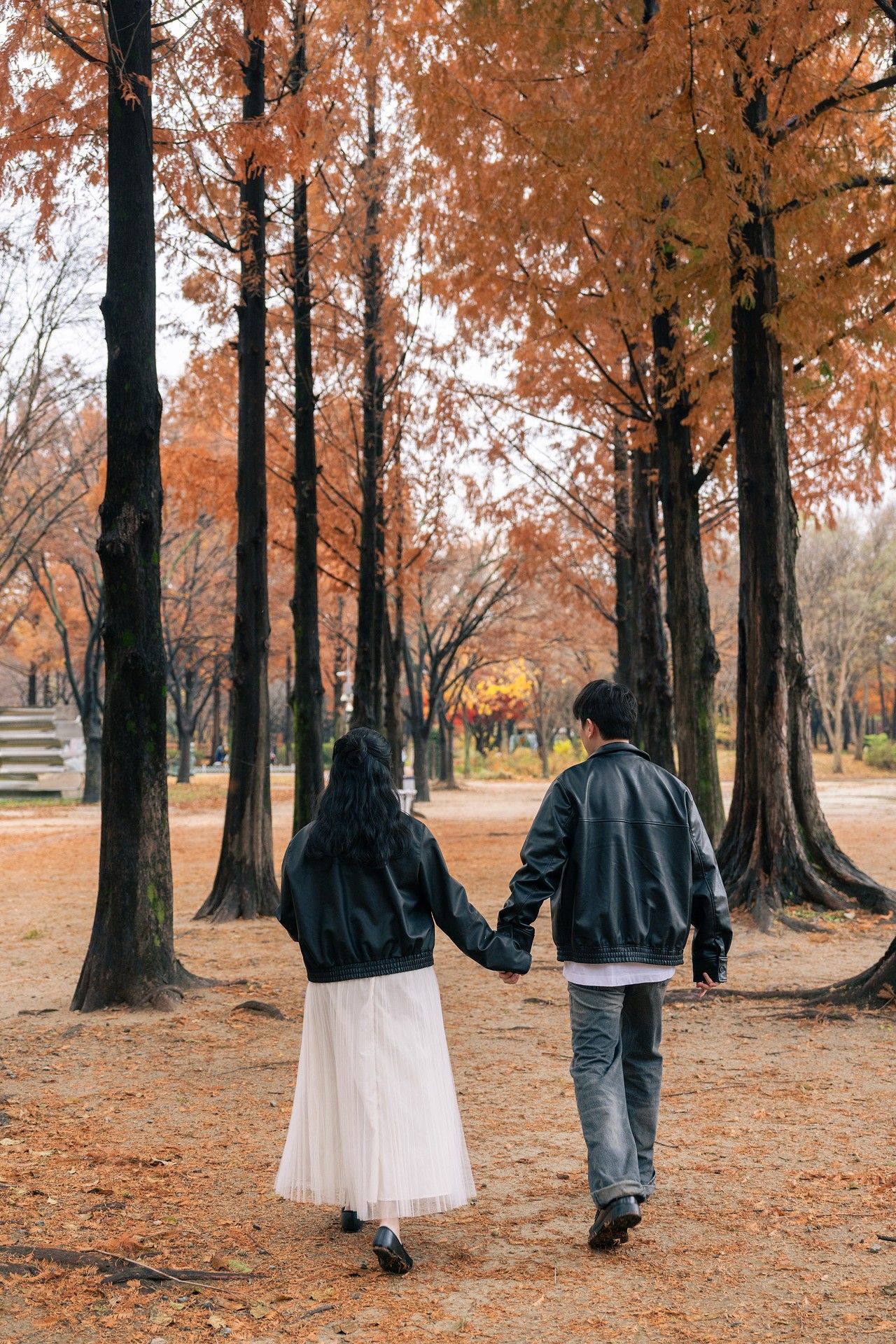 A couple walks through a forest path covered in fallen autumn leaves as orange foliage decorates the trees above.
