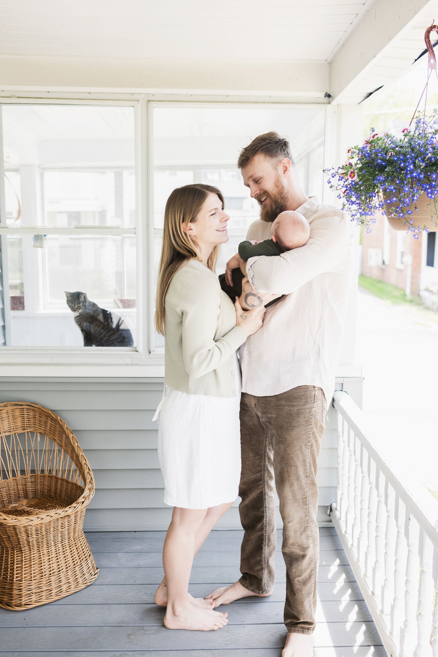 A happy couple holds newborn son swaddled in forest green on porch as their cat peers through window for a newborn shoot in Southern, Maine.